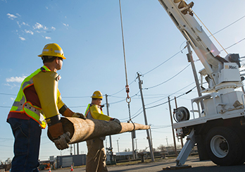 Two workers in hard hats guiding a utility pole that is being hoisted by a crane